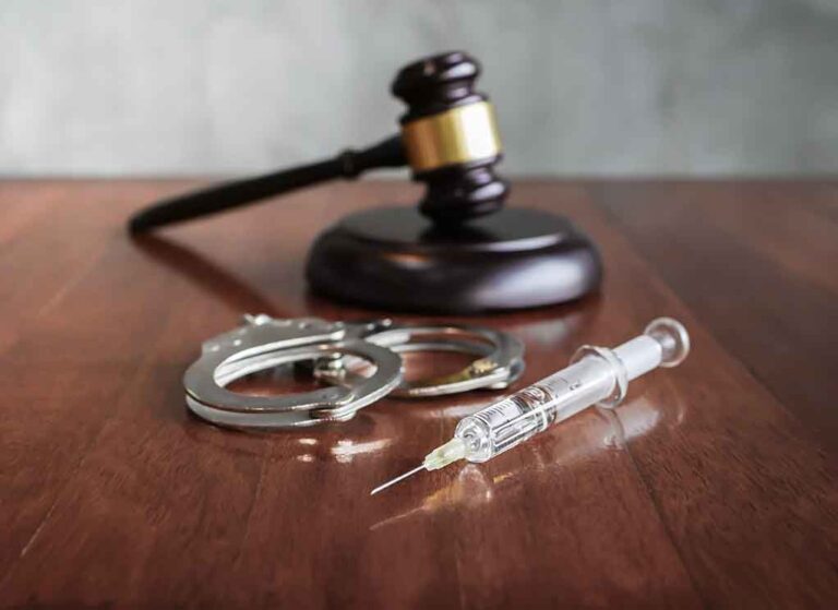 Gavel, handcuffs, and syringe on wooden table.