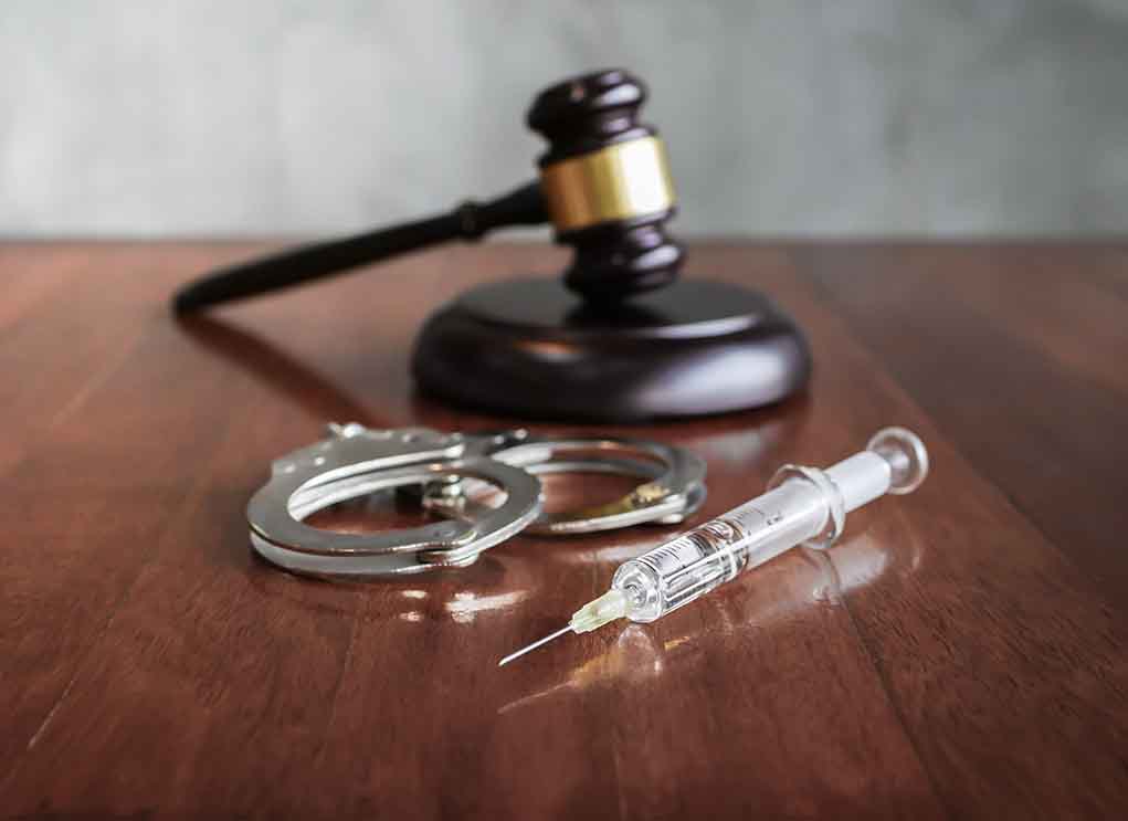 Gavel, handcuffs, and syringe on wooden table.