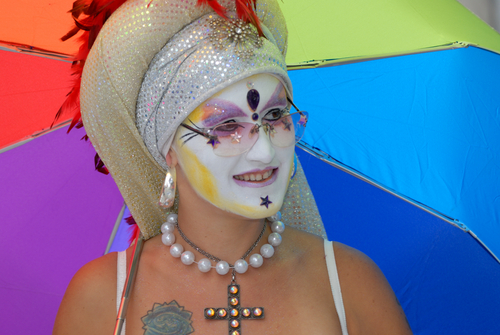 Person in colorful attire and makeup holding rainbow umbrella.
