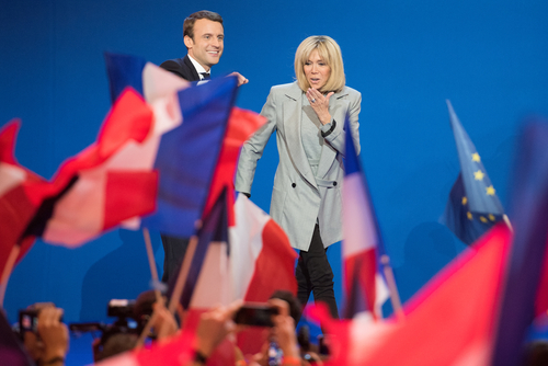 Emmanuel and Brigitte Macron on stage with waving French flags in the background