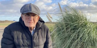 Elderly man in a black jacket and cap sitting outdoors near tall grass